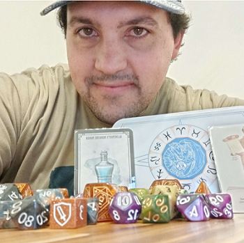 A white man wearing a hat holds TTRPG items and sits at a table with multicolored polyhedral dice