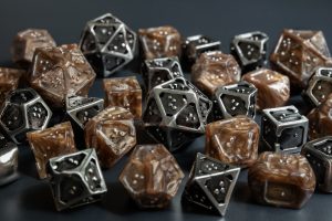 Close up view of a pile with multiple braille polyhedral dice. The sets are metallic bronze acrylic with dark pearl swirls and silver hollow metal with black antiqued finish. Both braille dice sets use a single character per face on all dice. The edges around all dice faces are raised with one border having a small divot in the center to act as an orientation marker, signifying the bottom of a braille cell. The other edges around the face set are flatter and thicker to provide increased stability when reading.