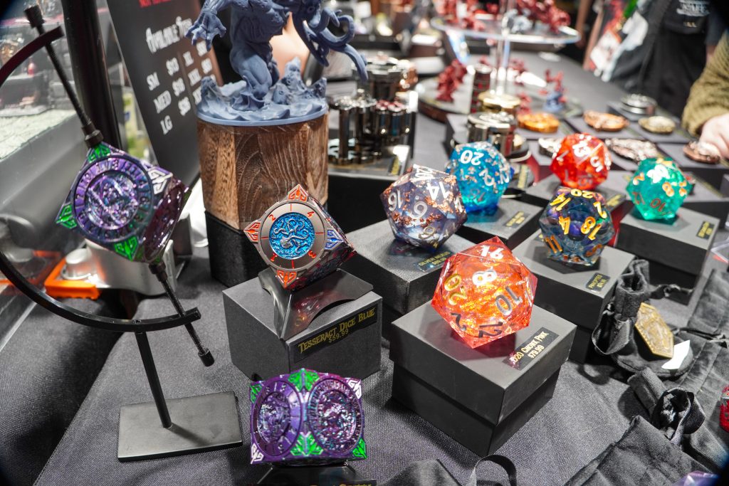 An assortment of giant, colorful D20 dice, including transparent and glitter-infused designs, displayed on stands at the Lethal Shadows booth.