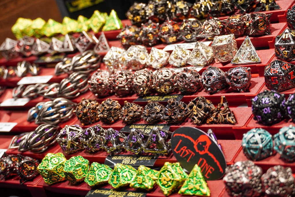 A collection of ornate, metal dice sets in various finishes and designs, labeled with product names and prices at the Lethal Shadows booth.