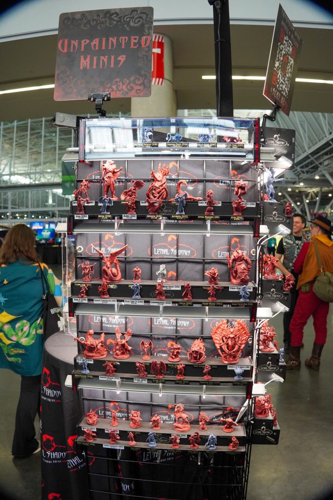 A display of unpainted miniatures arranged on shelves at the Lethal Shadows booth, labeled “Unpainted Minis,” with a variety of detailed fantasy and monster figures.