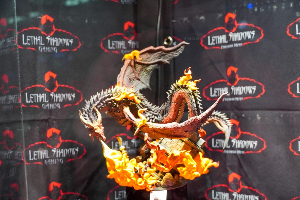 A close-up of an intricately sculpted dragon breathing fire, painted in fiery reds and oranges, displayed at the Lethal Shadows booth against a branded backdrop.