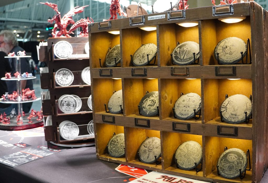 A display of character-themed coasters, each engraved with fantasy class icons like Paladin, Wizard, and Samurai, housed in a rustic wooden shelf.