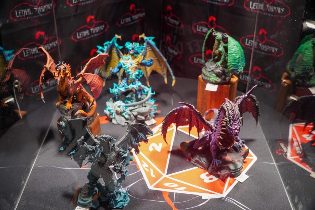 A showcase of large, painted dragon miniatures and other fantastical creatures arranged on a D20-themed mat at the Lethal Shadows booth.