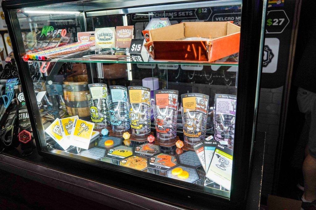 Display case at the RollACrit booth showcasing Heroes of Barcadia-themed glassware, dice, and game cards, along with character cards for game expansion packs.
