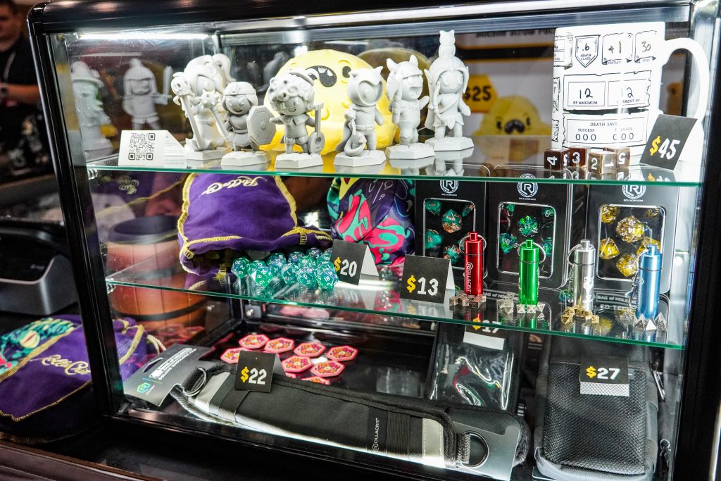 Display case at the RollACrit booth featuring unpainted miniatures, velvet dice bags, metal dice containers, and RPG-themed coffee mugs with character sheet designs.