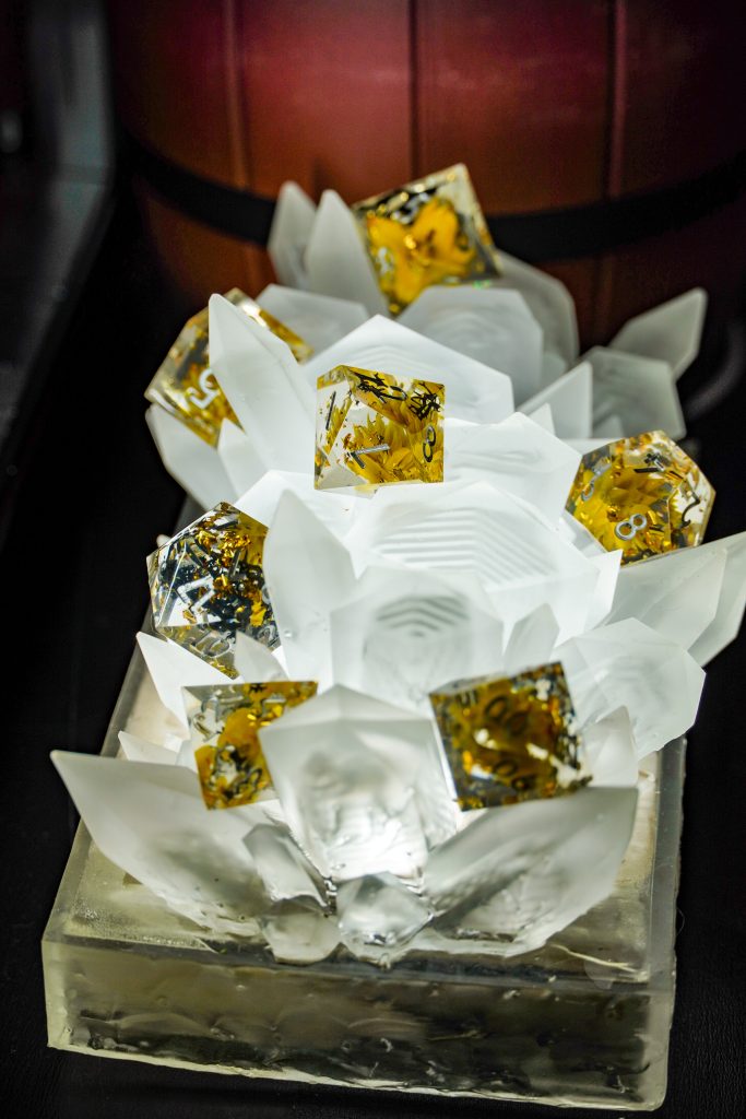 Close-up of a decorative dice display featuring translucent white resin spikes holding gold-accented dice with glitter inside, set against a wooden barrel backdrop.