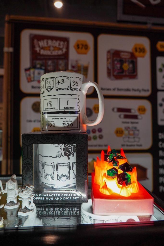 RPG-themed coffee mug designed like a character sheet, placed alongside a dice set displayed in a flame-themed holder, at the RollACrit booth.